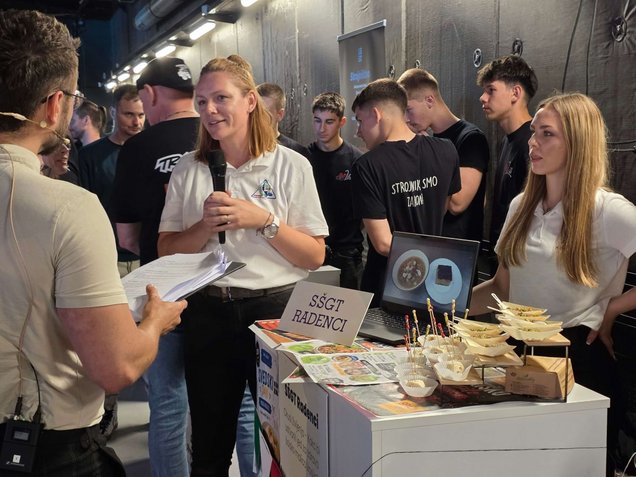 Zaključni dogodek Mojstri prihodnosti 3. junija 2026 v Ljubljani: mentorica skupaj z dijakinjami srednje strokovne šole v pogovoru z voditeljem prireditve na razstavnem prostoru predstavlja jedi, ki so jih pripravile kot odgovor na praktični izziv.